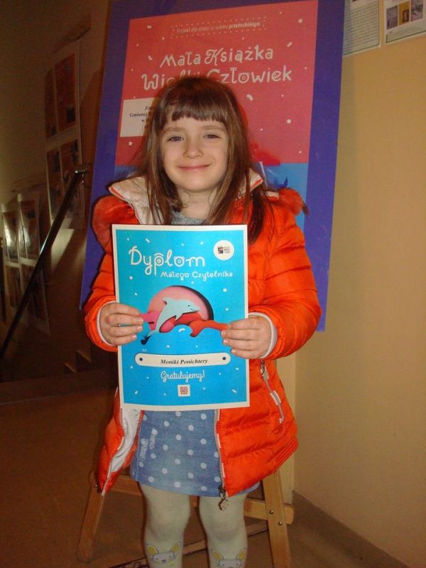 Zdjęcie przedstawia kilkuletnią dziewczynkę, trzymającą w rękach Dyplom Małego Czytelnika
